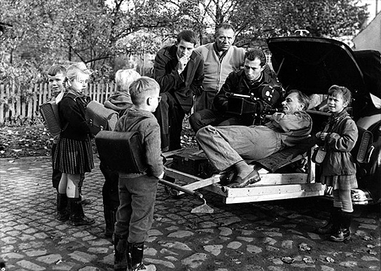 Regisseur Winfried Junge (links), Kamera-Assistent Hans Eberhard Leupold (3.v.l.), Kameramann Hans Dumke (im Auto liegend) bei Dreharbeiten mit Kindern der 1. Klasse in Golzow, 1961 ("Lebensläufe. Die Geschichte der Kinder von Golzow in einzelnen Portraits", 1981); Quelle: BArch