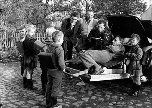 Regisseur Winfried Junge (links), Kamera-Assistent Hans Eberhard Leupold (3.v.l.), Kameramann Hans Dumke (im Auto liegend) bei Dreharbeiten mit Kindern der 1. Klasse in Golzow, 1961 ("Lebensläufe. Die Geschichte der Kinder von Golzow in einzelnen Portraits", 1981); Quelle: BArch